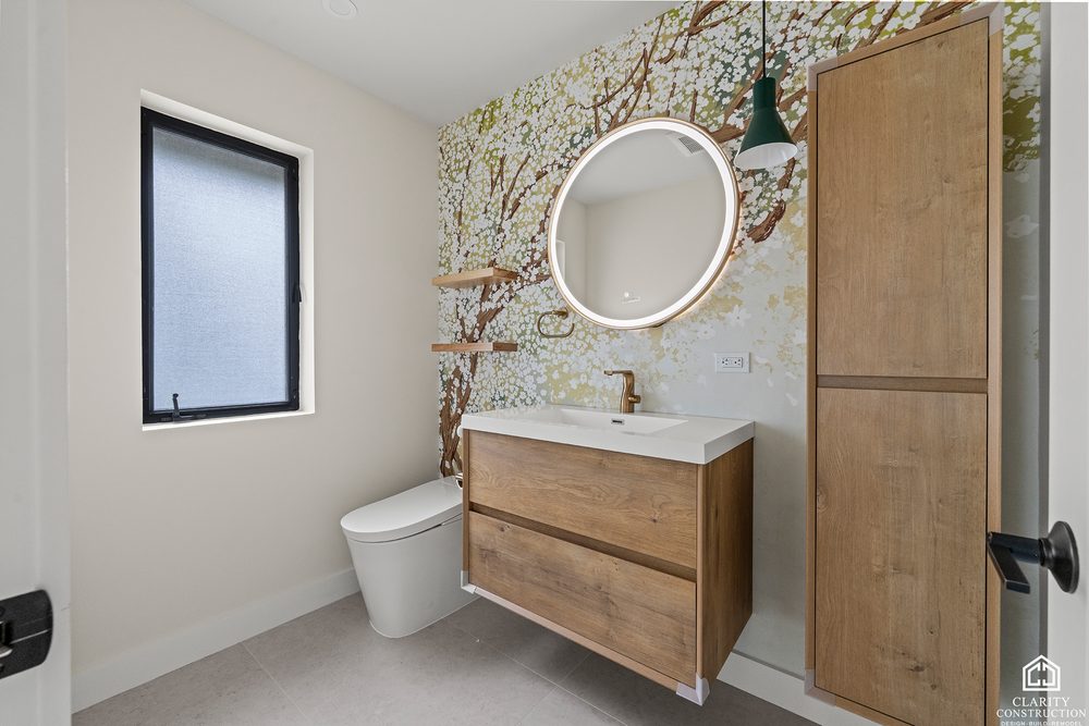 Bathroom with a floral mural wall, an oak vanity and a round backlit mirror